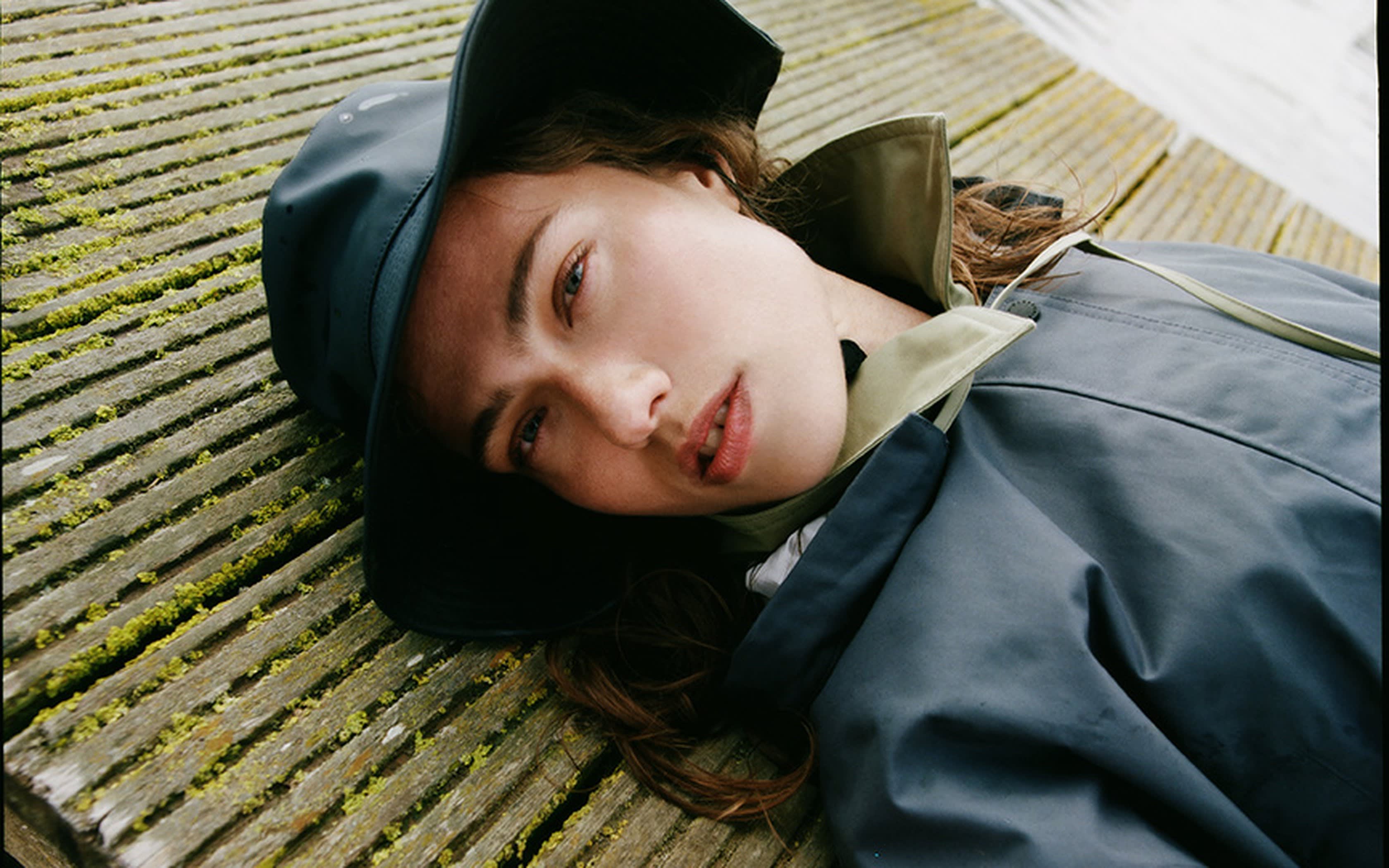 Femme avec chapeau de pluie noir et veste bicolore allongée sur un ponton.