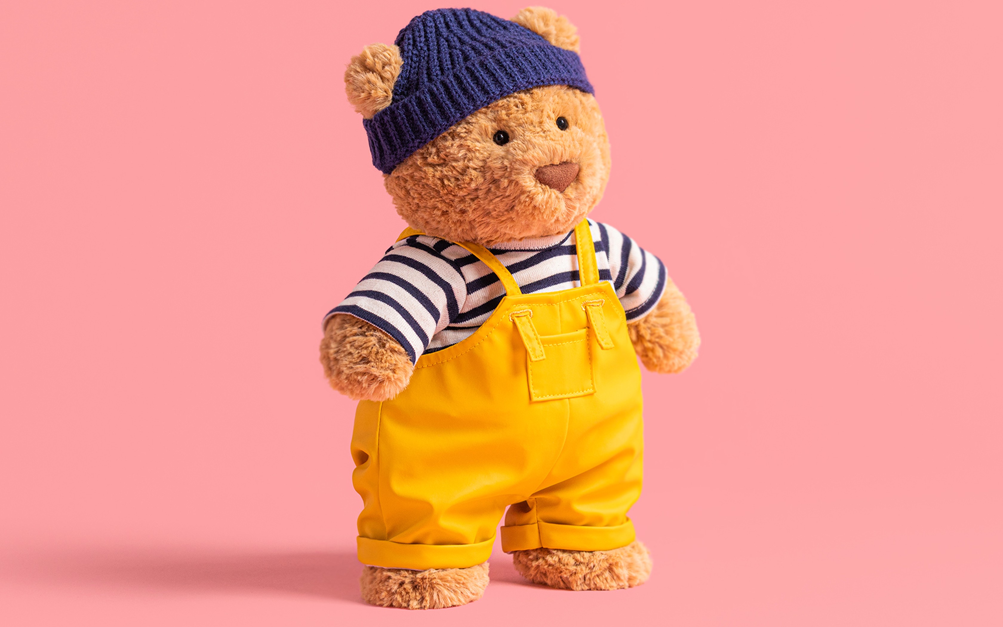 Ours en peluche avec bonnet bleu, t-shirt rayé et salopette jaune sur fond rose.