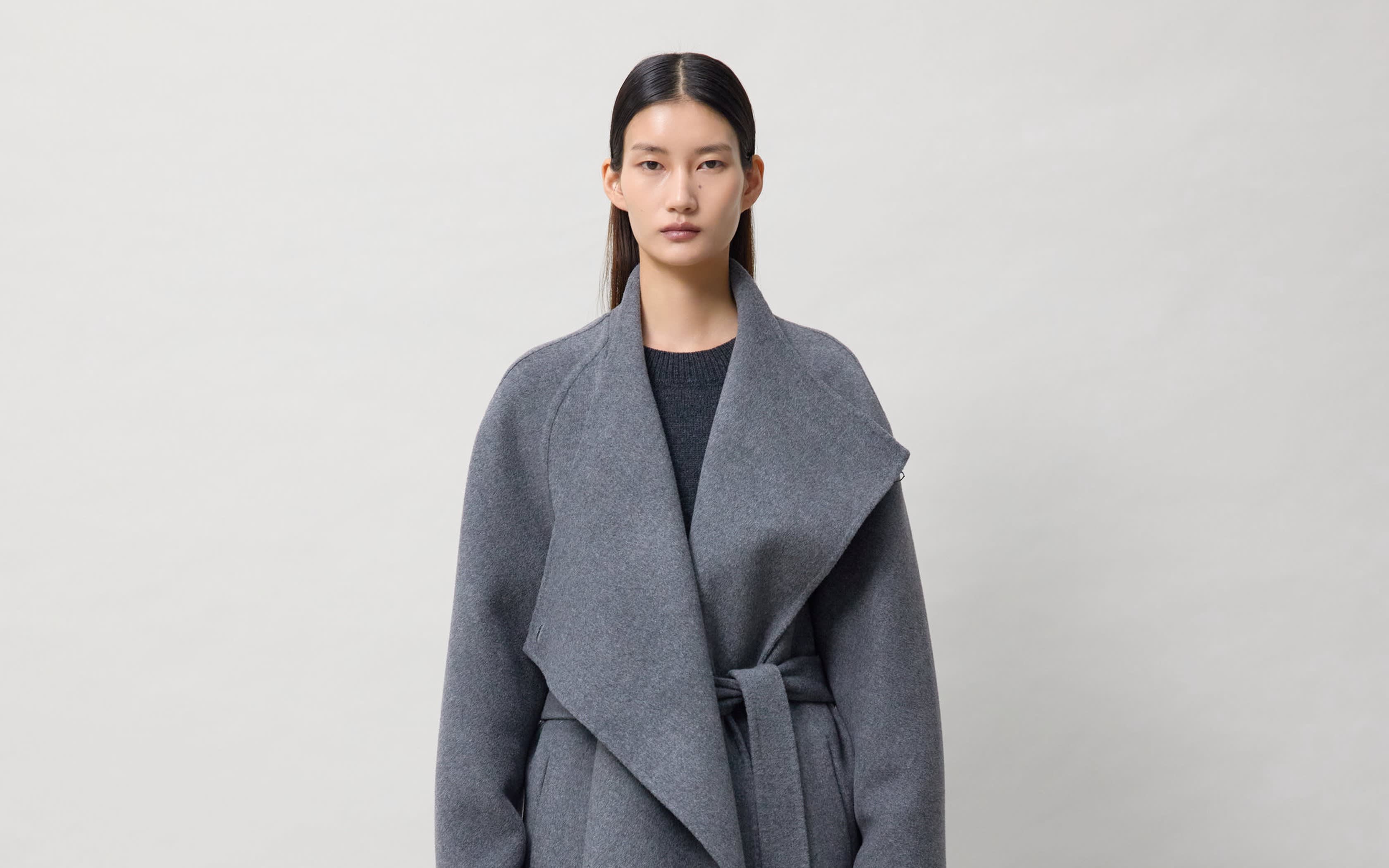 Femme portant un long manteau gris ceinturé sur fond neutre.