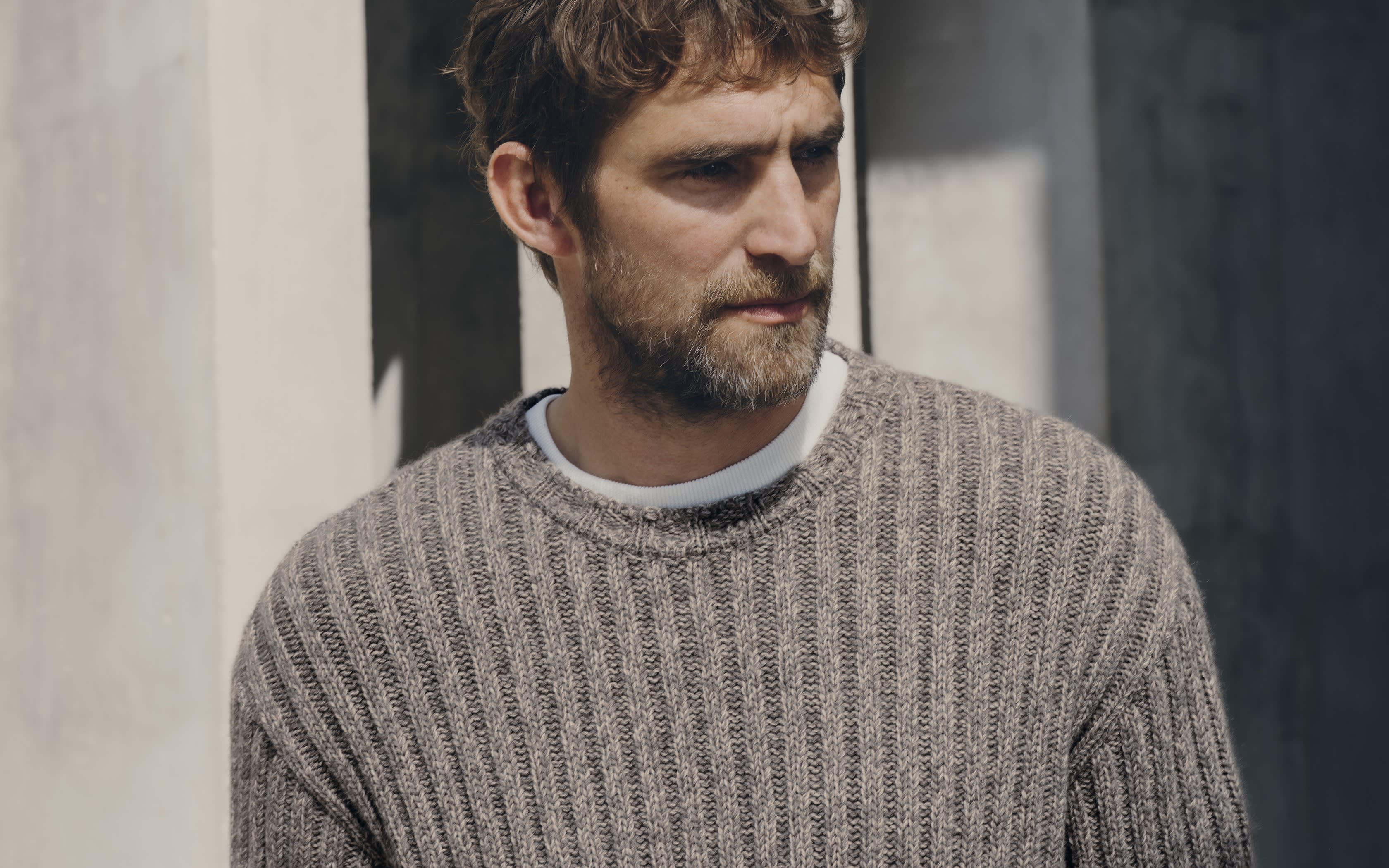 Ein bärtiger Mann in einem braun-grauen Rippstrick-Pullover vor einer Betonwand.