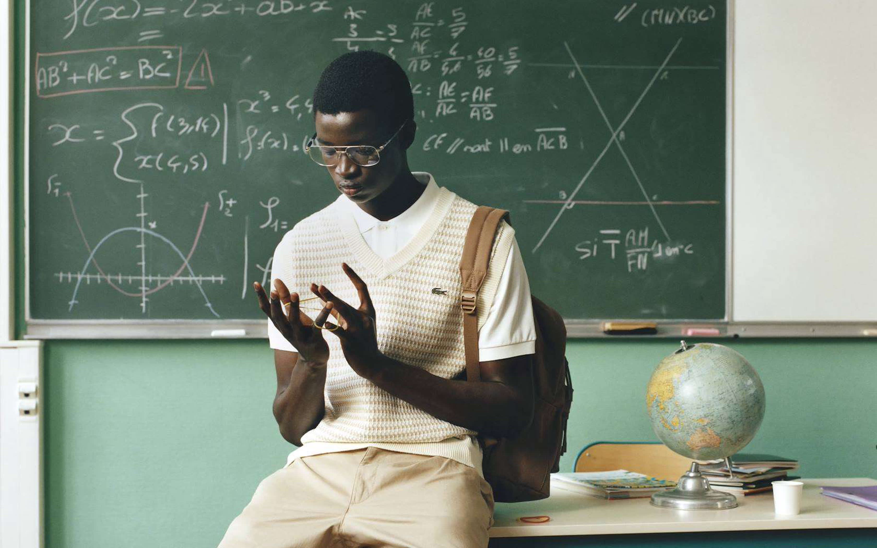 Junger Mann im Klassenzimmer mit Strickpullover und Brille sitzt auf dem Lehrertisch.